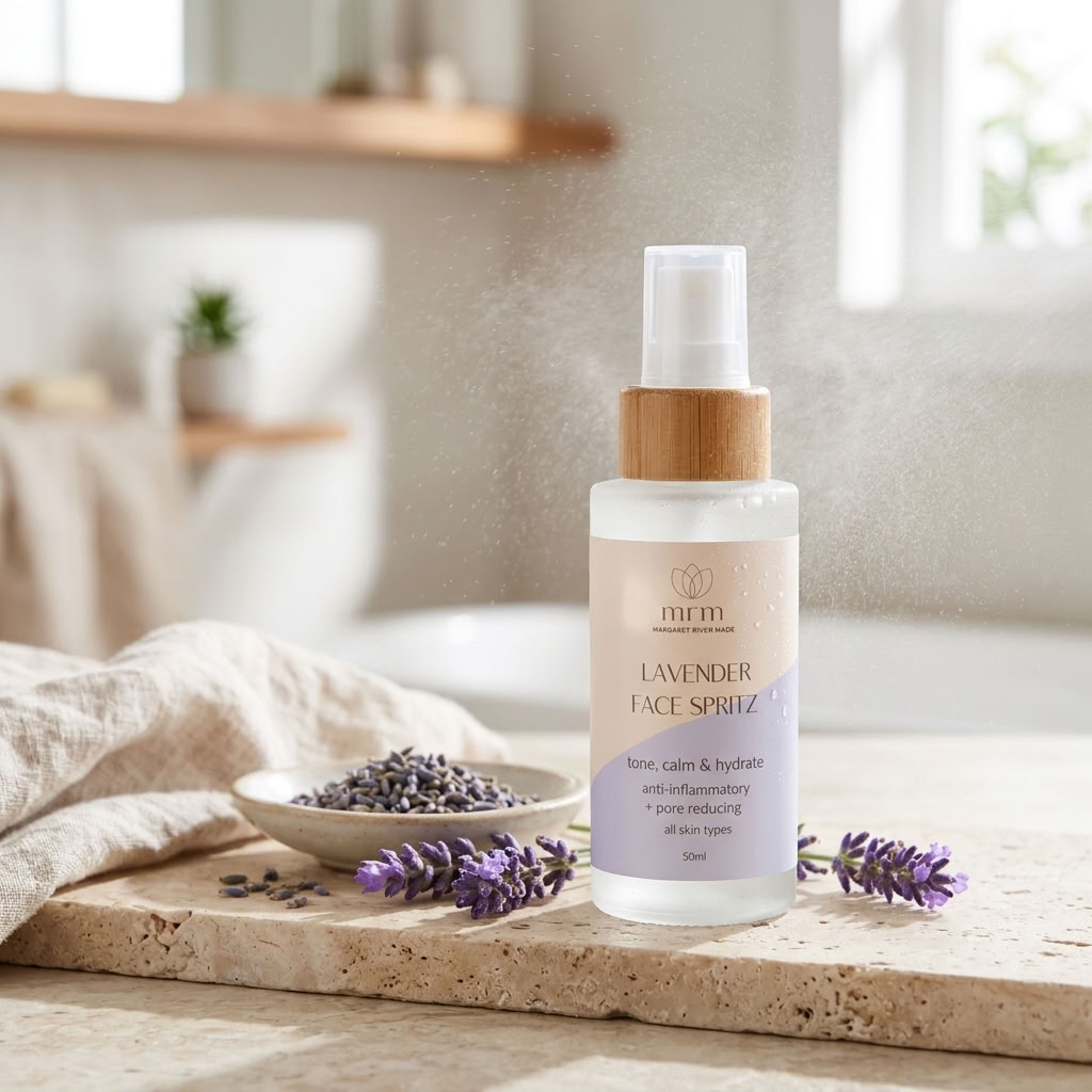 A bottle of MFM Lavender Face Spray stands on a stone surface with lavender sprigs and a small dish of dried lavender in a softly lit bathroom-like setting. The product bottle is frosted with a bamboo-style pump and clear spray top.