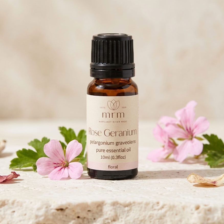 A 10ml bottle of mrm Rose Geranium essential oil, styled on a textured stone surface with pink flowers and green leaves against a soft beige background.