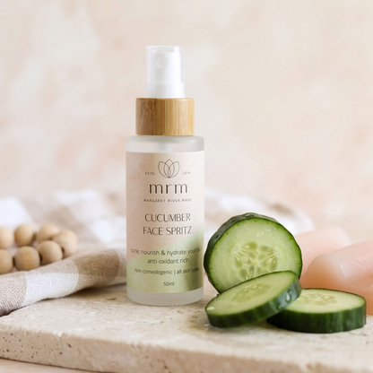 A 50ml bottle of MRM Cucumber Face Spritz with a bamboo cap, styled on a stone surface with cucumber slices, a folded towel, and a pink soap bar in the background.