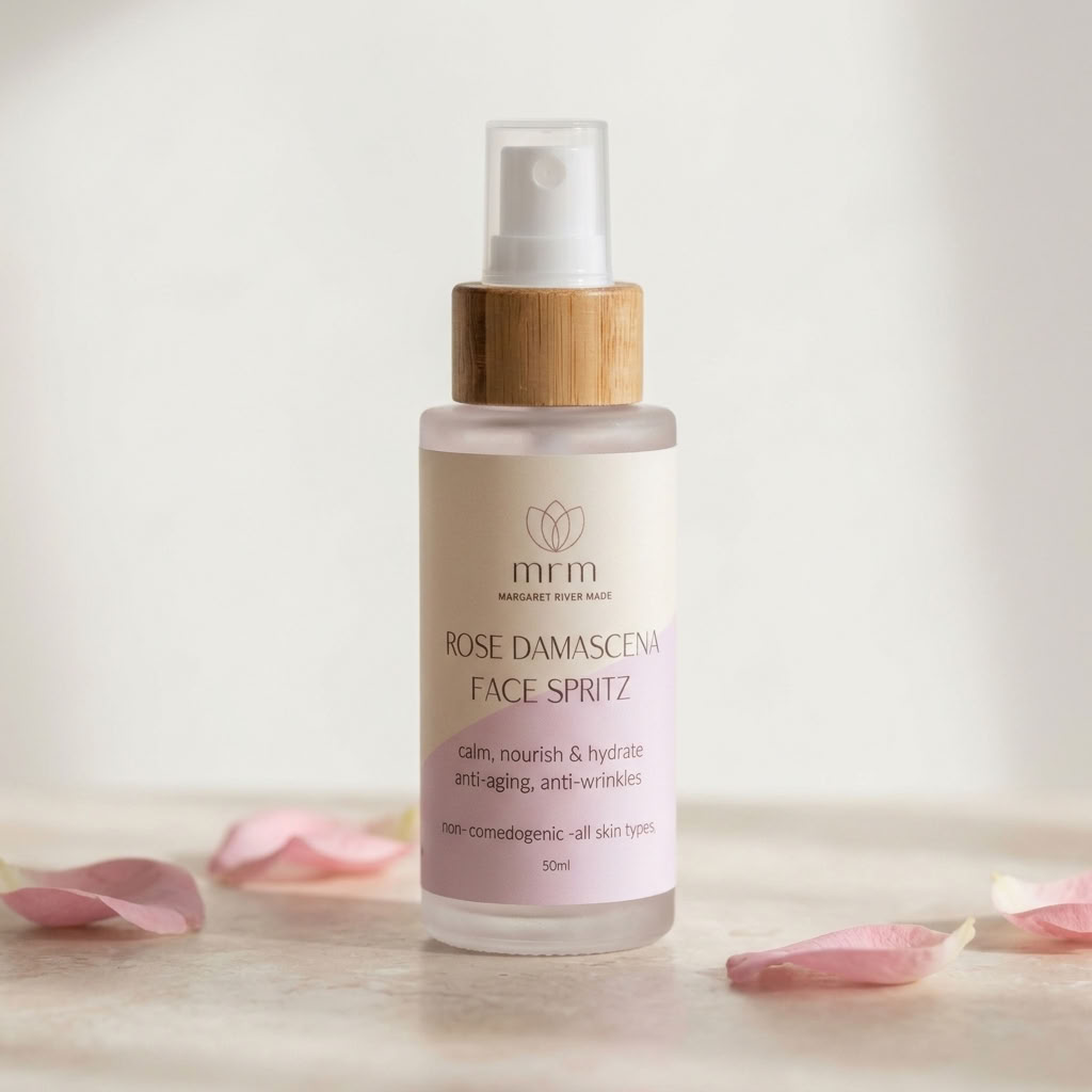 A 50ml bottle of mrm Rose Damascena Face Spritz with a bamboo cap, set on a light surface with scattered pink rose petals against a soft, warm off-white background.