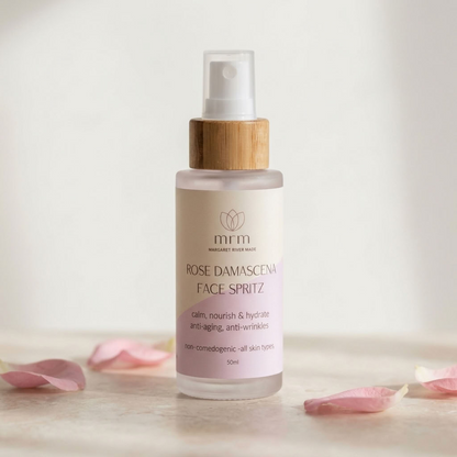 A 50ml bottle of mrm Rose Damascena Face Spritz with a bamboo cap, set on a light surface with scattered pink rose petals against a soft, warm off-white background.