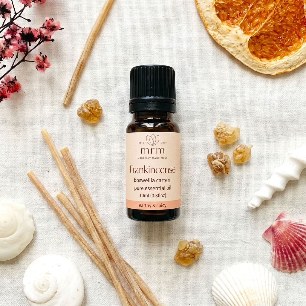 A 10ml bottle of MRM Frankincense pure essential oil, styled in a flat-lay arrangement with natural ingredients like frankincense buds, cinnamon sticks, dried orange slices, and seashells on a textured cream fabric background.