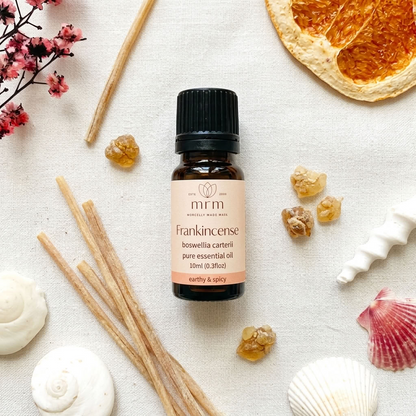 A 10ml bottle of MRM Frankincense pure essential oil, styled in a flat-lay arrangement with natural ingredients like frankincense buds, cinnamon sticks, dried orange slices, and seashells on a textured cream fabric background.