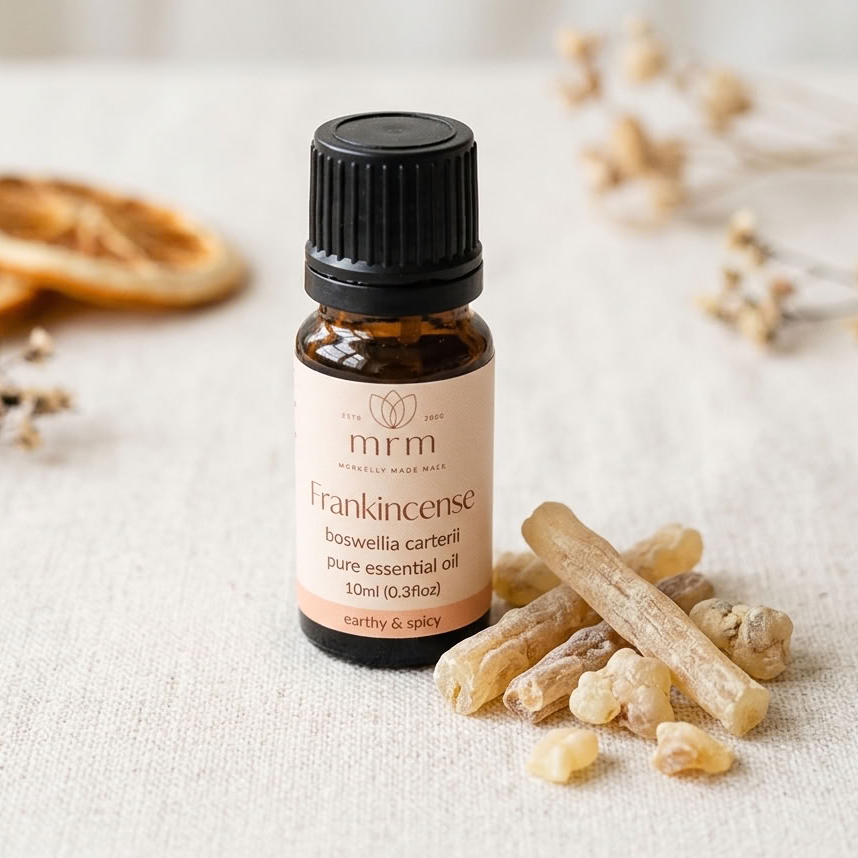 A small amber glass bottle of MRM Frankincense pure essential oil with a black cap, set on a textured cream linen surface with dried orange slices and incense sticks as props.