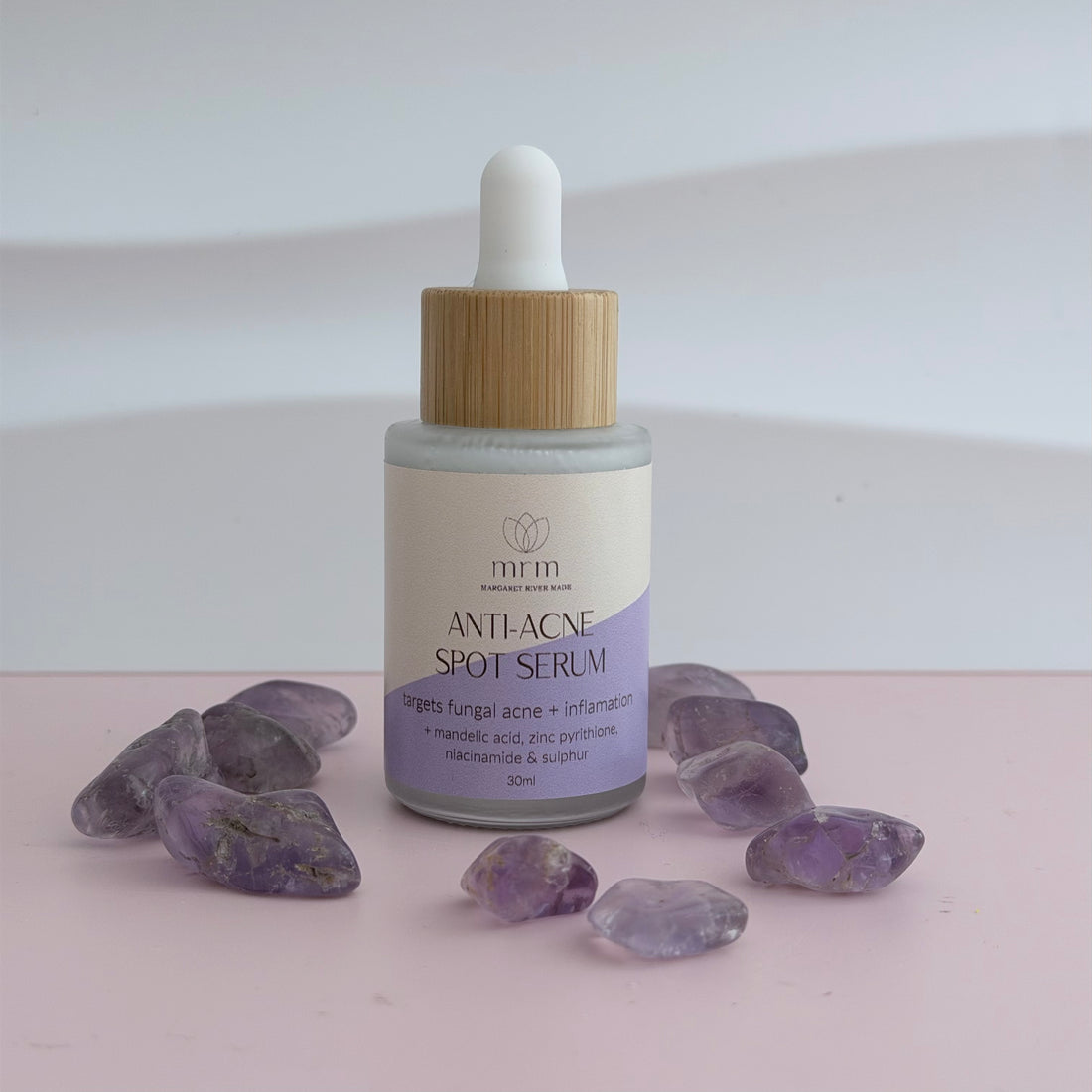 Anti-acne spot serum bottle with wooden dropper on a light surface with purple stones.